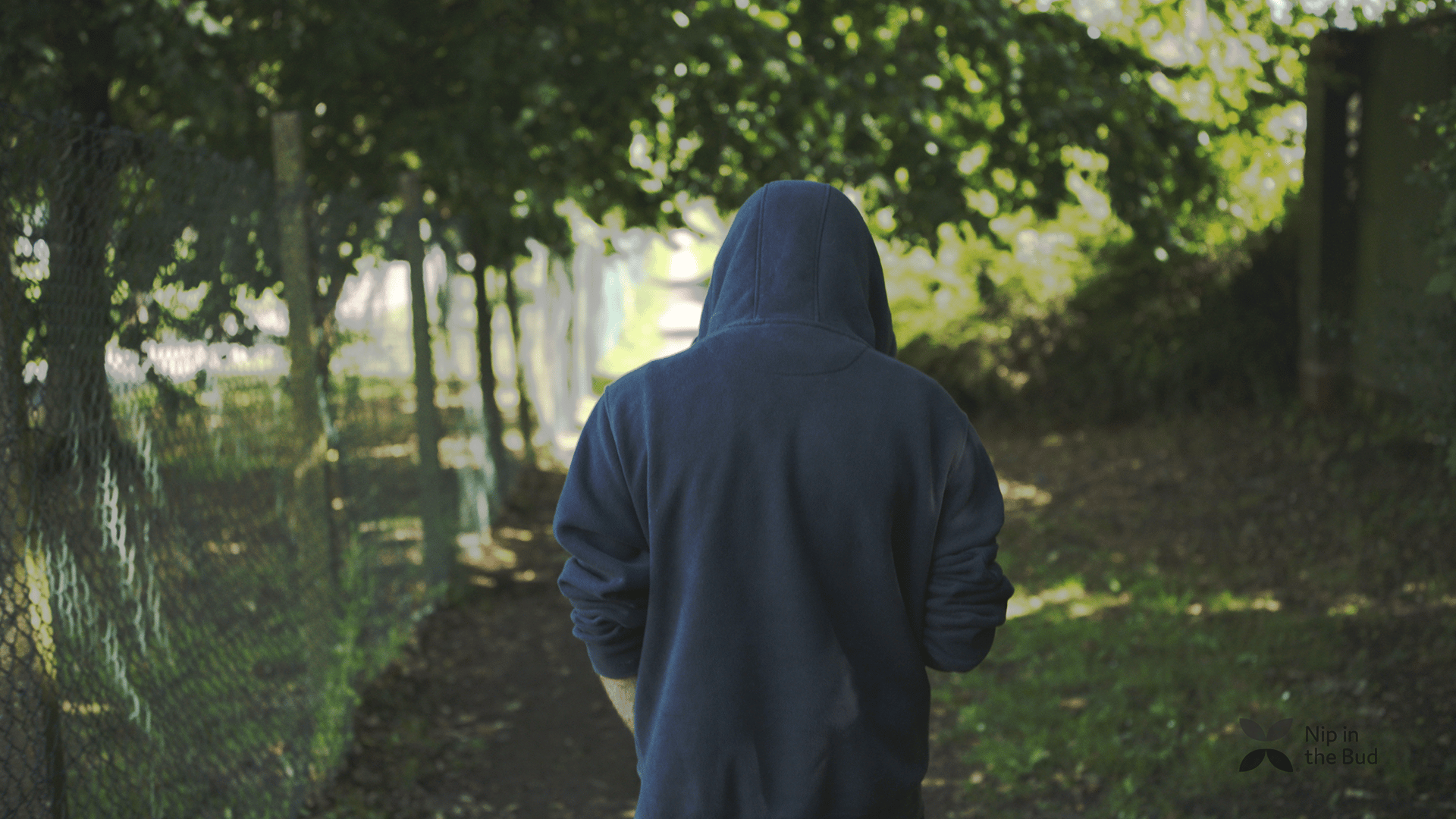 A young man wearing a hoodie is walking away through a tree lined path.